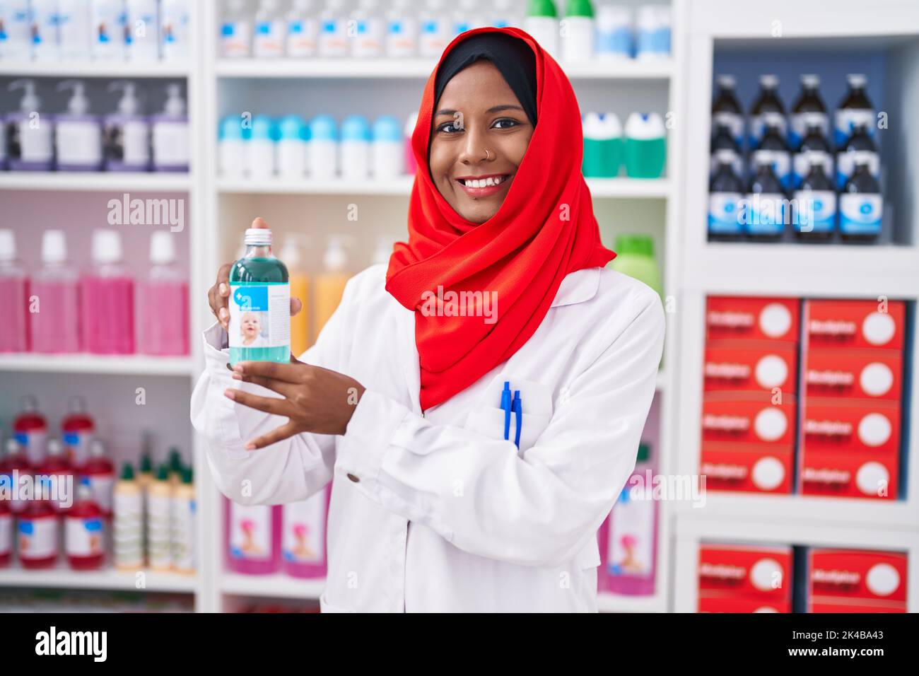 Jeune femme arabe travaillant à la pharmacie pharmacie pharmacie de pharmacie holding sirop regardant positif et heureux debout et souriant avec un sourire confiant montrant des dents Banque D'Images