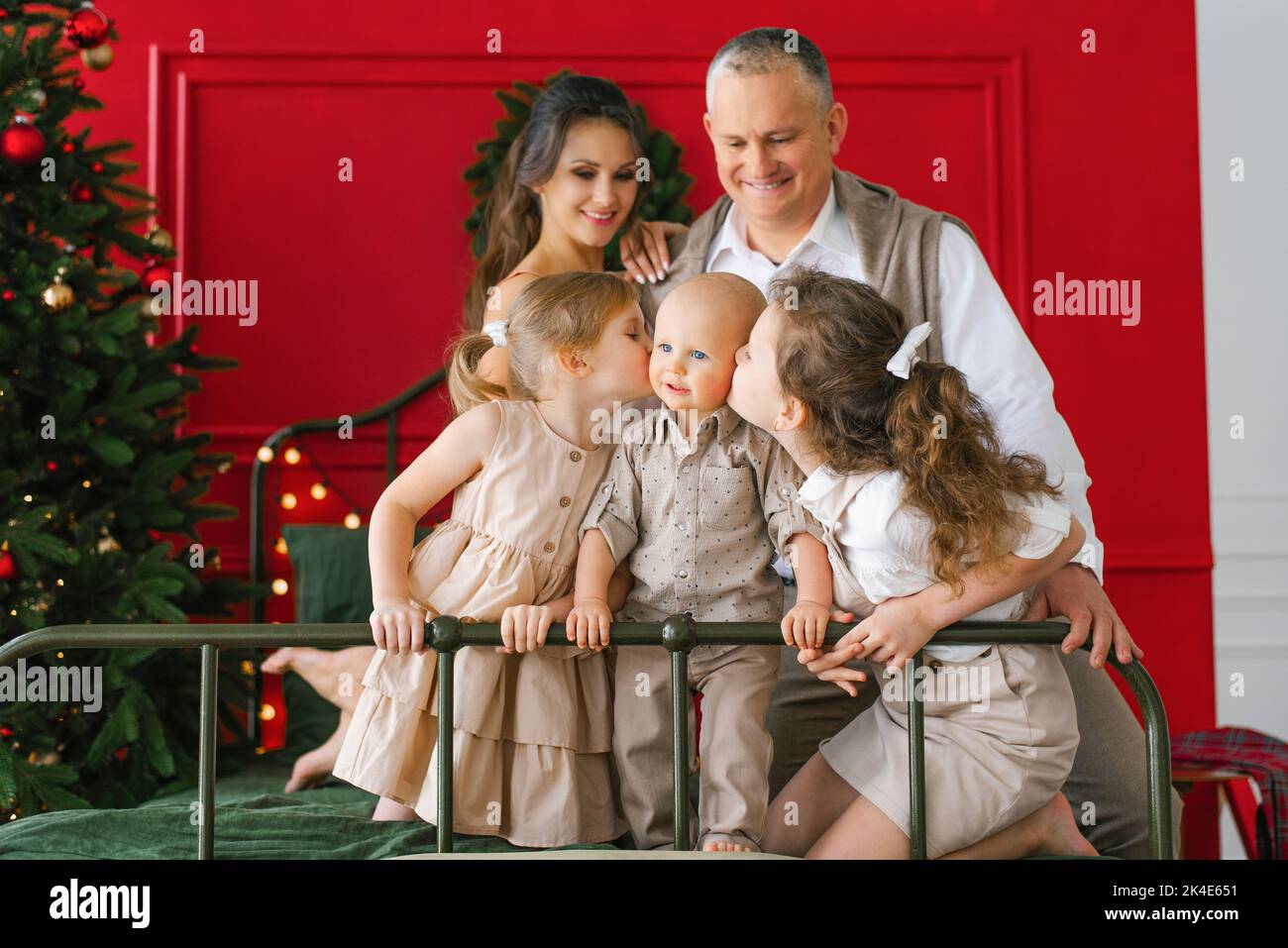Une famille élégante avec trois enfants est assise sur un lit près d'un arbre de Noël. Des vacances de Noël confortables. Les sœurs embrassent leur frère, les parents regardent le Banque D'Images