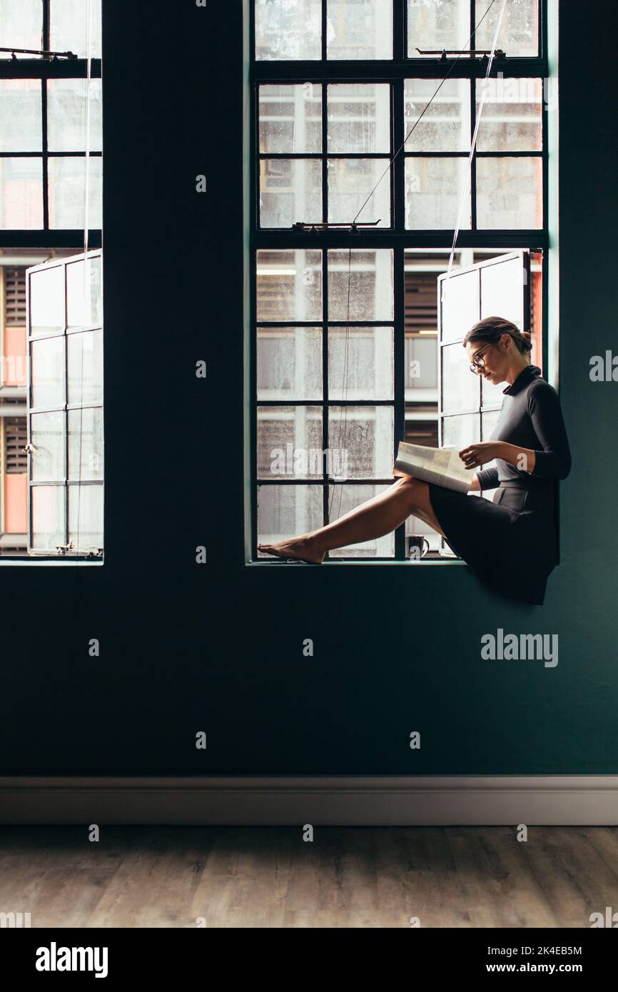 Vue latérale d'une femme asiatique assise sur le rebord de la fenêtre et lisant un livre. Photo verticale d'une femme lisant un magazine près de sa fenêtre de bureau. Banque D'Images