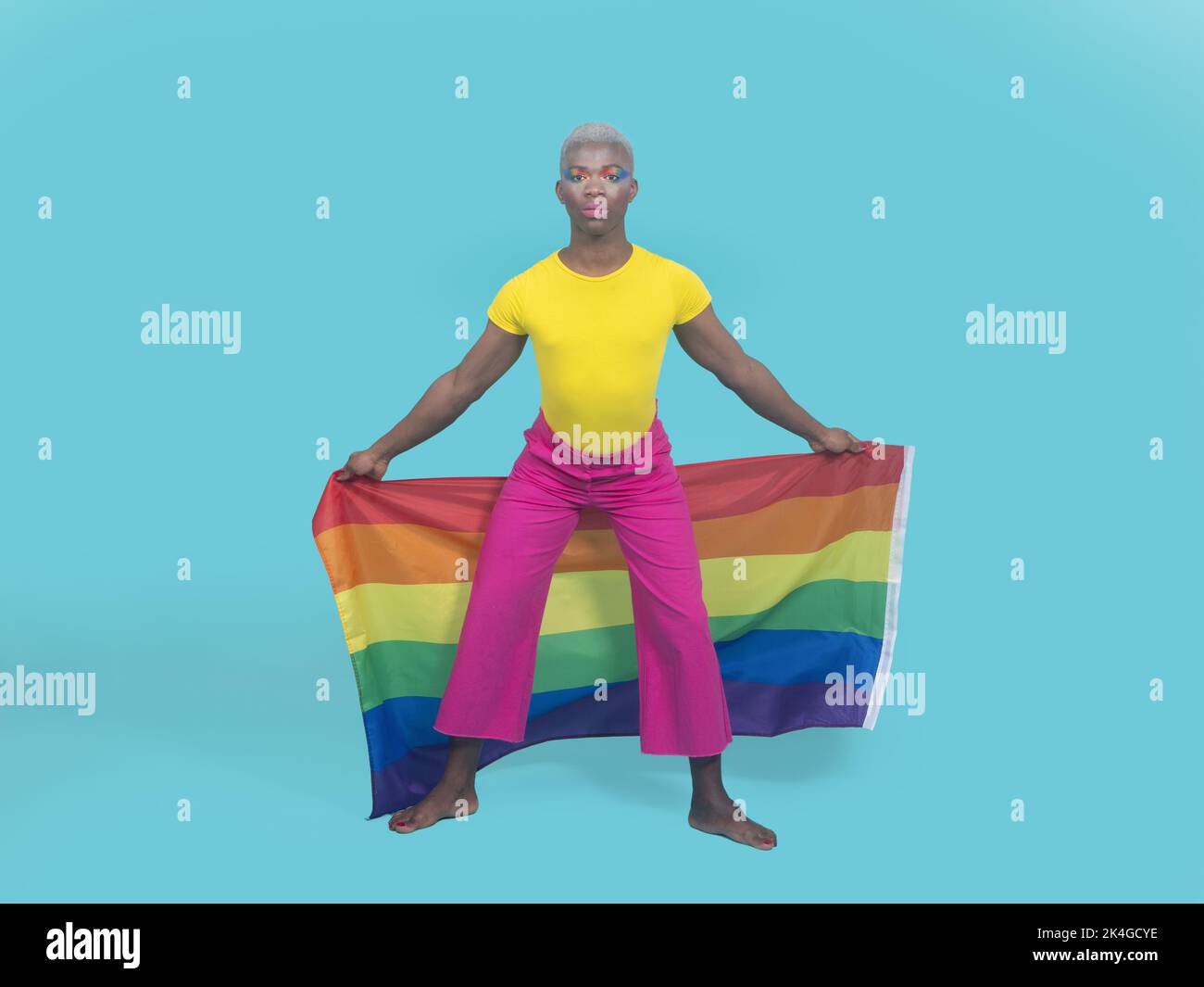 Tout le corps pieds nus noir gay dans des vêtements colorés regardant l'appareil photo et en agitant le drapeau arc-en-ciel derrière les jambes lors de l'événement de fierté LGBT contre le dos turquoise Banque D'Images