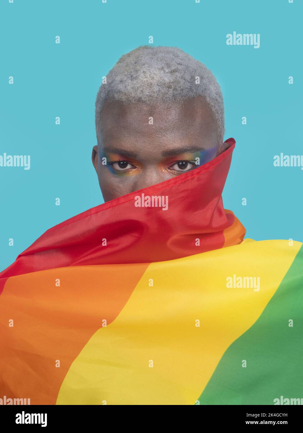 Gay noir avec maquillage et cheveux blonds couvrant le visage avec drapeau arc-en-ciel sur fond turquoise Banque D'Images