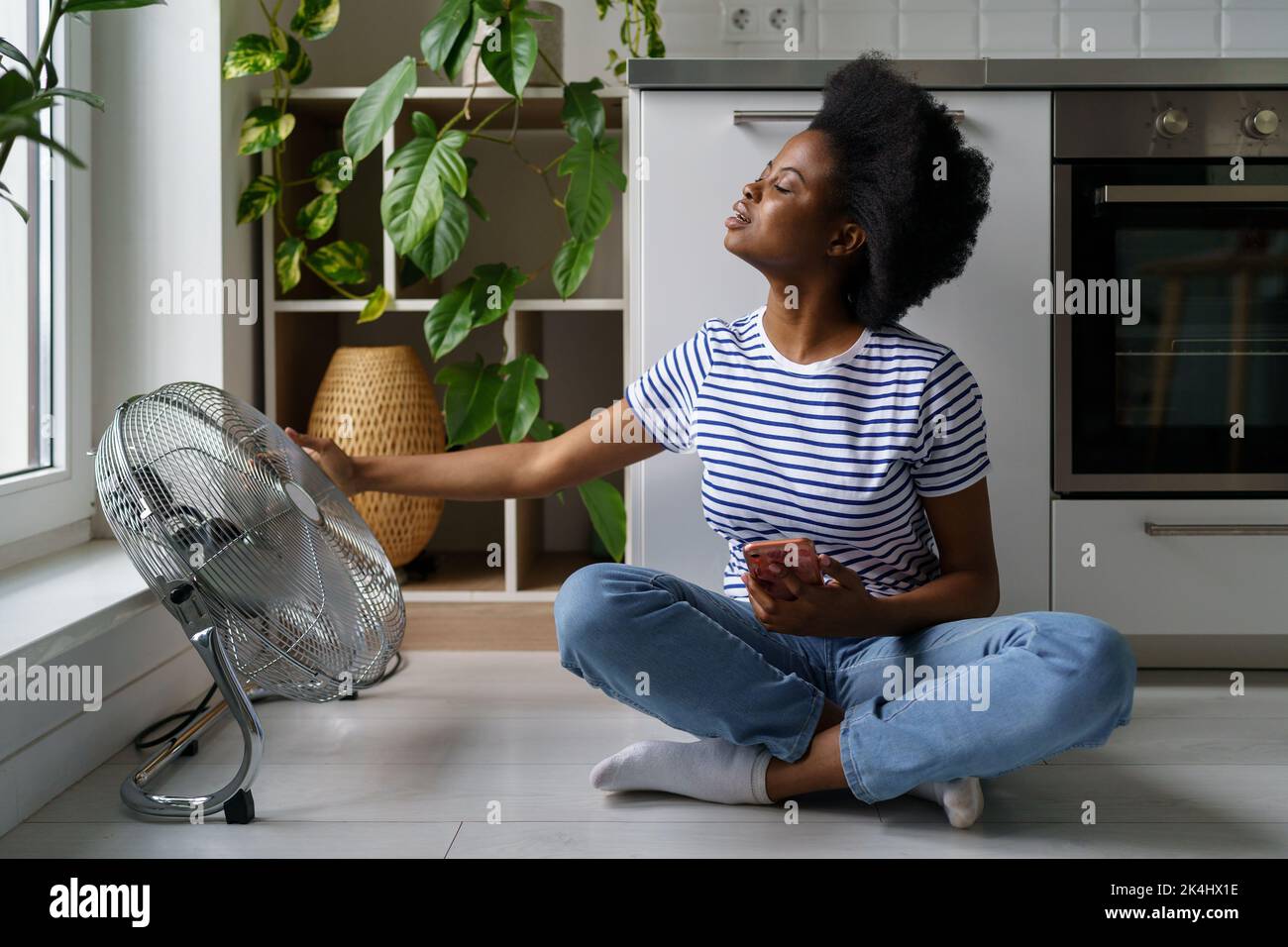 Une jeune femme africaine est assise sur le sol avec un téléphone et attrape les courants d'air émanant du ventilateur électrique Banque D'Images