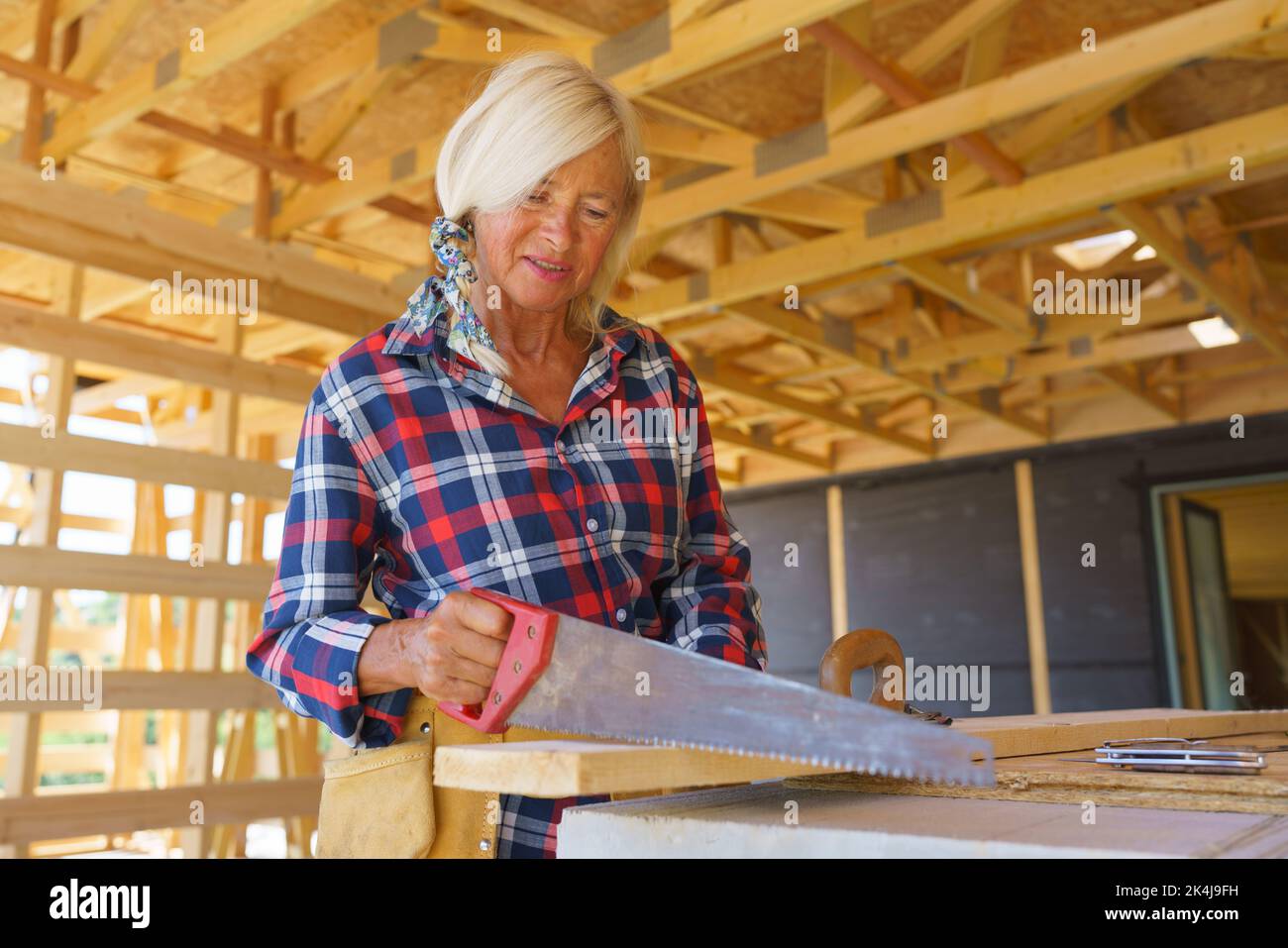 Femme âgée sciant une planche à bois à l'intérieur de son éco-maison en bois écologique durable inachevé. Concept de femmes et de personnes âgées actives et indépendantes, éco Banque D'Images
