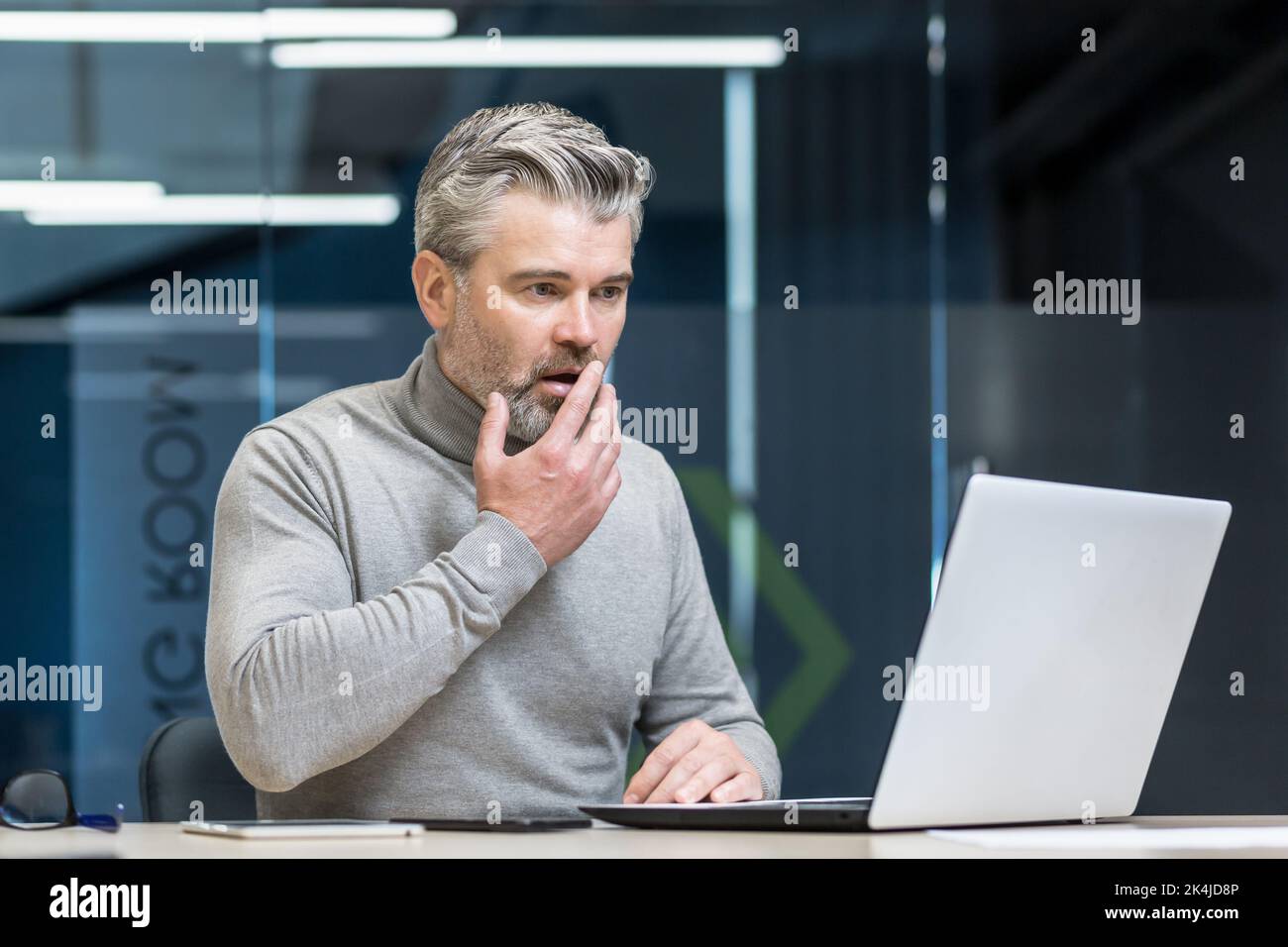 Un homme choqué, un homme d'affaires aux cheveux gris, assis au bureau, travaille avec un ordinateur portable. Il tient sa main à la bouche, surpris par ce qu'il a vu sur l'écran de l'ordinateur. Banque D'Images