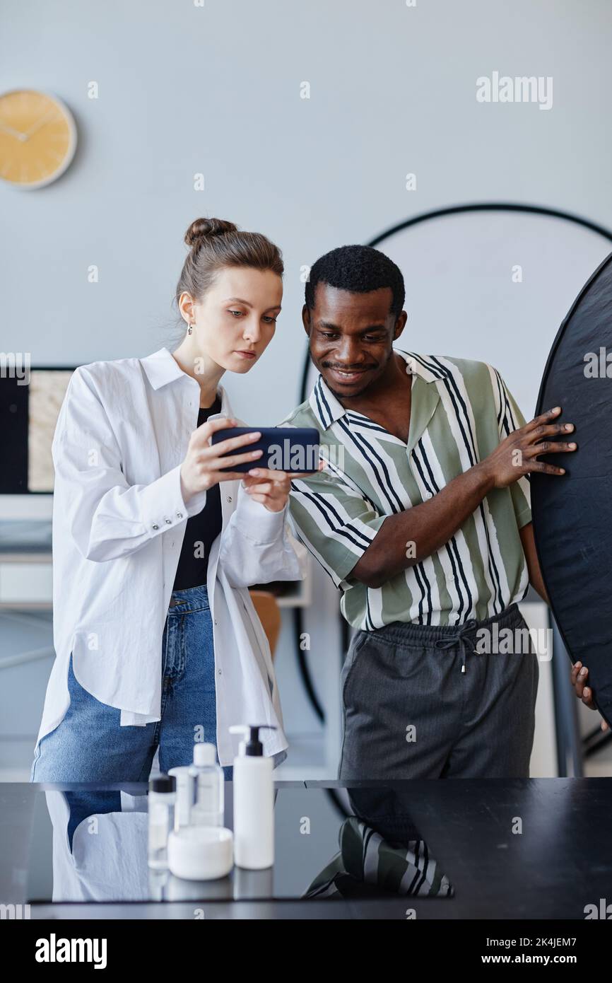Portrait vertical de deux jeunes photographes travaillant sur la photographie de produits en studio et utilisant un téléphone mobile pour les médias sociaux Banque D'Images