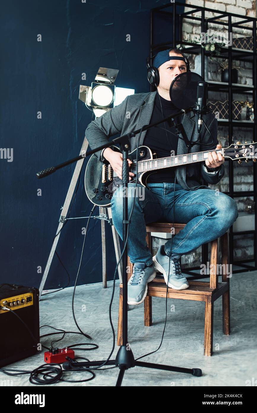 Gros plan d'un chanteur assis sur un tabouret dans un casque avec une guitare enregistrant une piste dans un studio à domicile Banque D'Images
