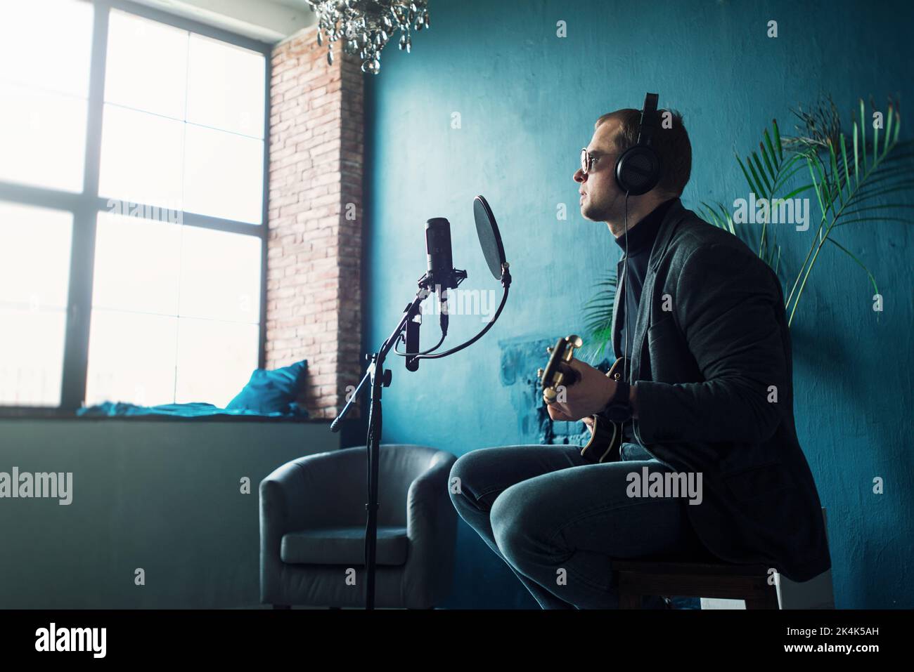 Gros plan d'un chanteur assis sur un tabouret dans un casque avec une guitare enregistrant une piste dans un studio à domicile Banque D'Images