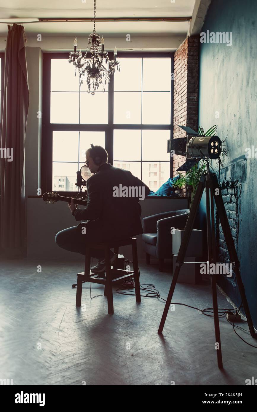 Homme chanteur assis sur un tabouret dans un casque avec une guitare enregistrant une piste dans un studio à domicile Banque D'Images