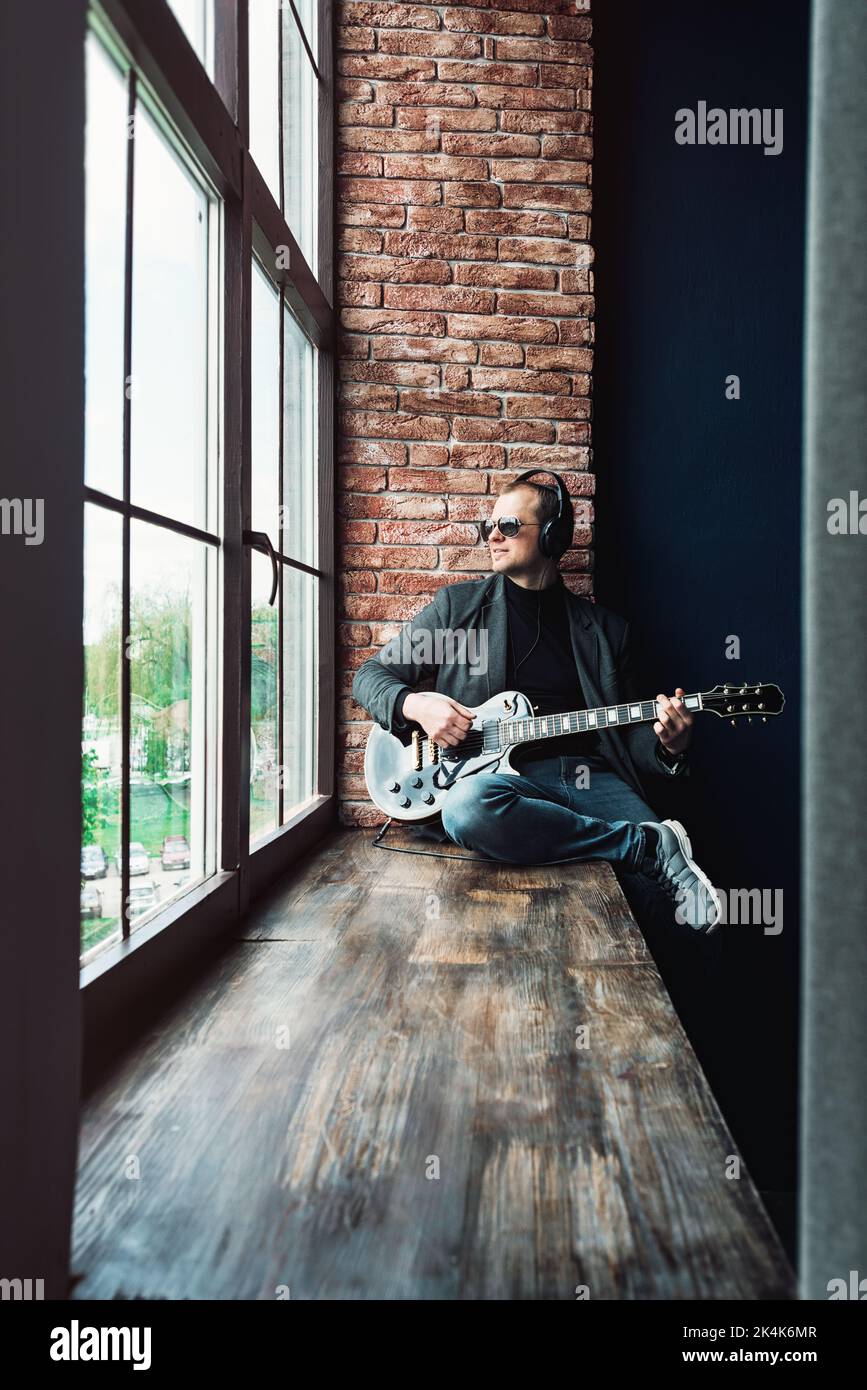 Homme chanteur assis sur un rebord de fenêtre dans un casque avec une guitare enregistrant une piste dans un studio à domicile Banque D'Images