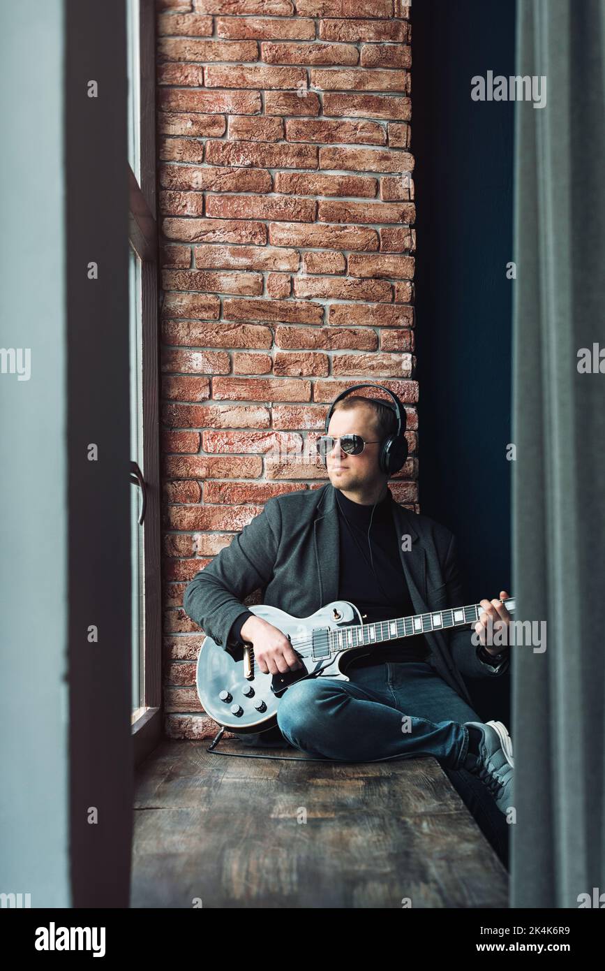 Homme chanteur assis sur un rebord de fenêtre dans un casque avec une guitare enregistrant une piste dans un studio à domicile Banque D'Images