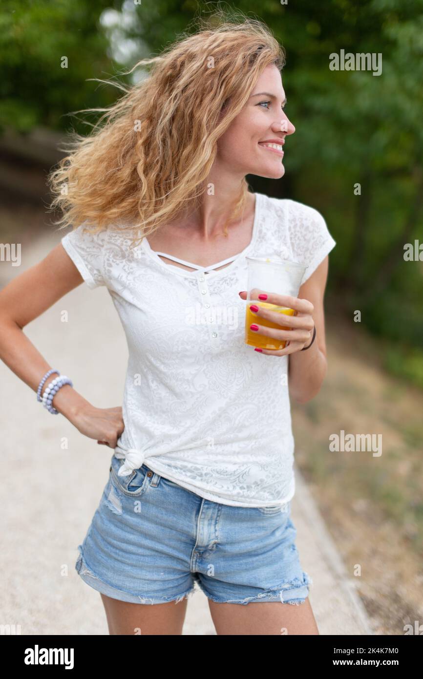 Bonne jeune caucasienne 30s femme tenant un verre de jus dans la nature et regardant loin Banque D'Images