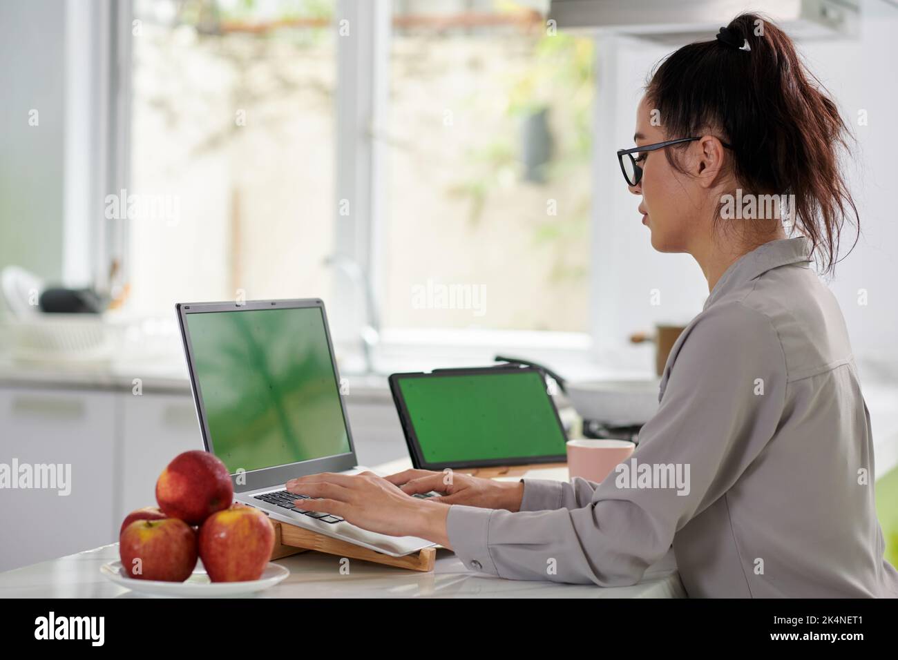 Vue latérale d'une jeune femme d'affaires moderne en tenue décontractée, dactylographiant sur un clavier d'ordinateur portable avec écran vert pendant le travail à domicile Banque D'Images