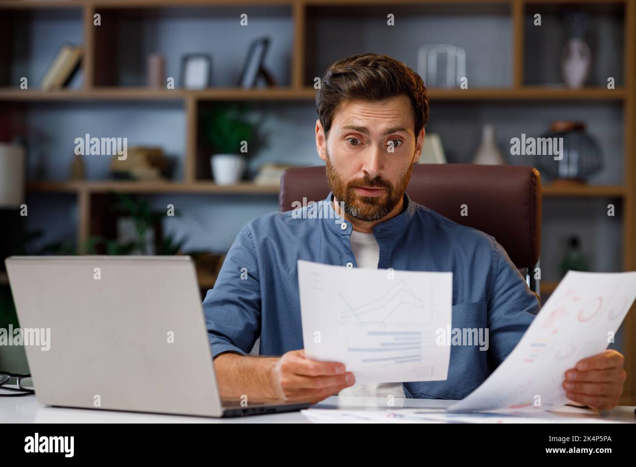 Beau homme travaillant sur un ordinateur portable tout en étant assis au bureau à domicile. Dans le stress, il examine les diagrammes de ventes, la crise financière sur la bourse. Mauvais Banque D'Images