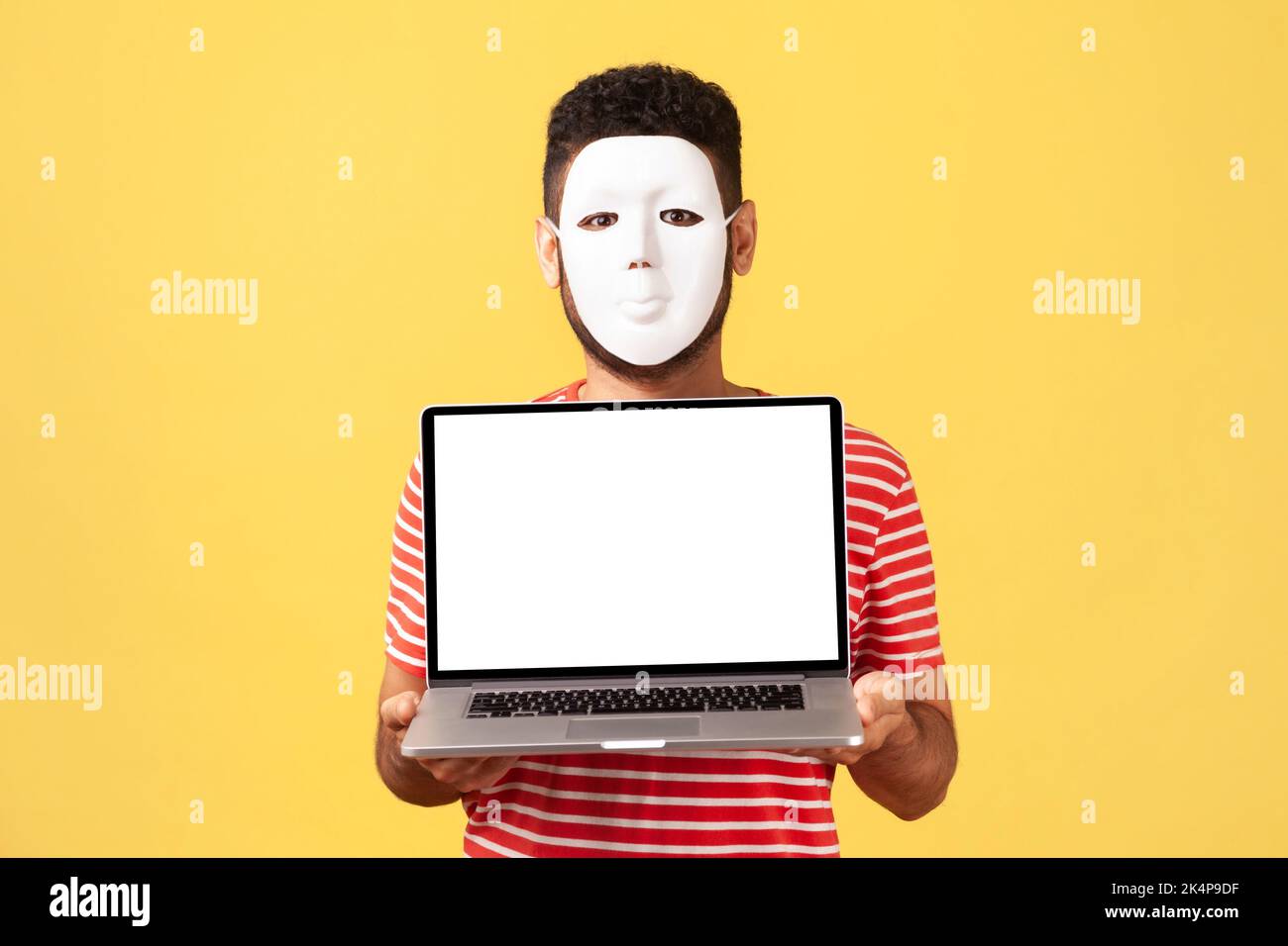 Portrait d'un homme anonyme inconnu en masque blanc cachant son vrai visage et sa personnalité, tenant un ordinateur portable avec écran vide, espace publicitaire, espace de copie. Studio d'intérieur tourné isolé sur fond jaune. Banque D'Images