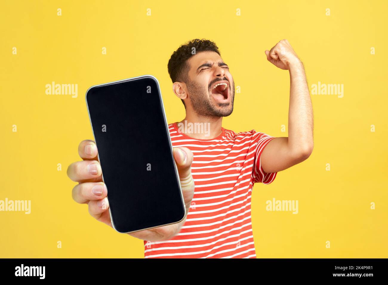 Très heureux homme joyeux avec la barbe portant un T-shirt rouge debout montrant un grand téléphone mobile avec un écran vide, célébrant son succès, gagnant. Studio d'intérieur isolé sur fond jaune. Banque D'Images Très heureux homme joyeux avec la barbe portant un T-shirt rouge debout montrant un grand téléphone mobile avec un écran vide, célébrant son succès, gagnant. Studio d'intérieur isolé sur fond jaune. Banque D'Images