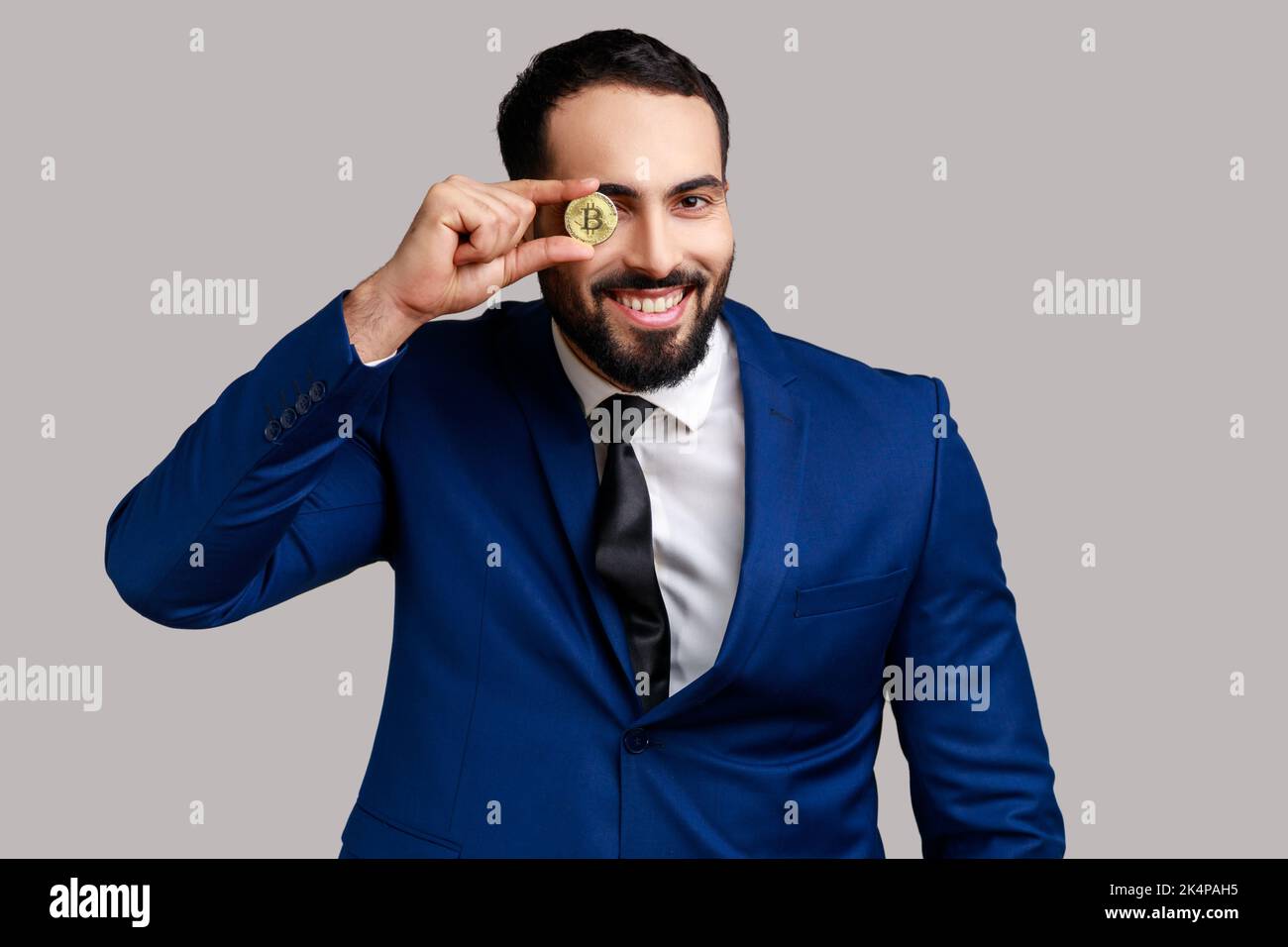 Portrait d'homme d'affaires barbu couvrant l'œil avec un bitcoin doré, regardant l'appareil photo avec un sourire éclatant, portant un costume de style officiel. Prise de vue en studio isolée sur fond gris. Banque D'Images