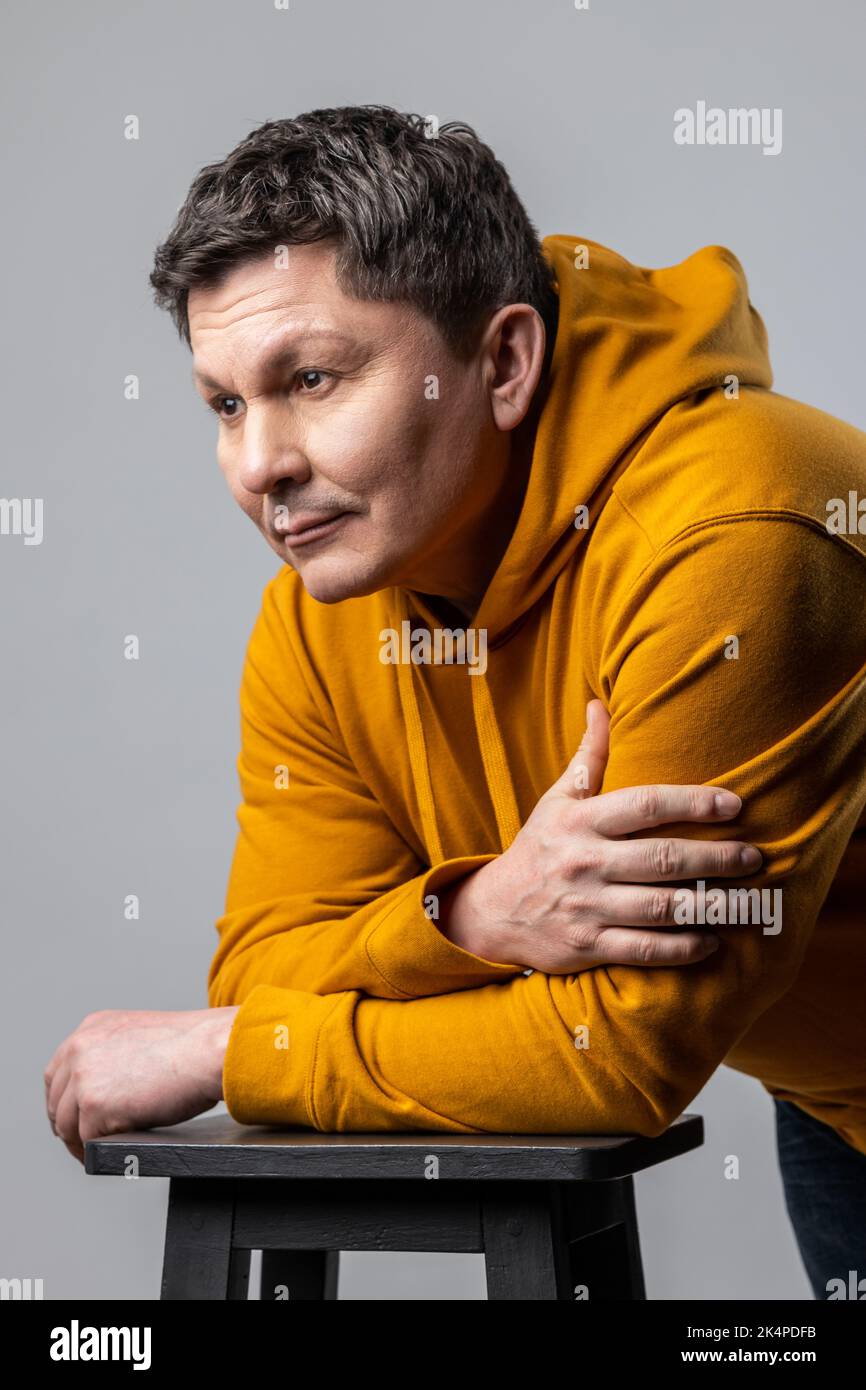 Portrait d'un beau homme d'âge moyen regardant pensif, pensif, penché sur la chaise et regardant loin, étant profondément dans les pensées, portant le sweat à capuche de style urbain. Prise de vue en studio isolée sur fond gris. Banque D'Images