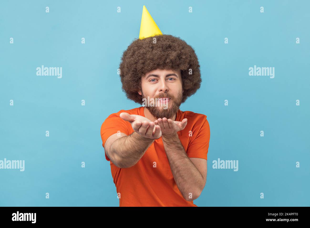 Portrait d'un homme romantique avec une coiffure afro en T-shirt orange et cône de fête envoyant des baisers d'air à ses invités, regardant l'appareil photo, célébrant l'anniversaire. Studio d'intérieur isolé sur fond bleu. Banque D'Images Portrait d'un homme romantique avec une coiffure afro en T-shirt orange et cône de fête envoyant des baisers d'air à ses invités, regardant l'appareil photo, célébrant l'anniversaire. Studio d'intérieur isolé sur fond bleu. Banque D'Images