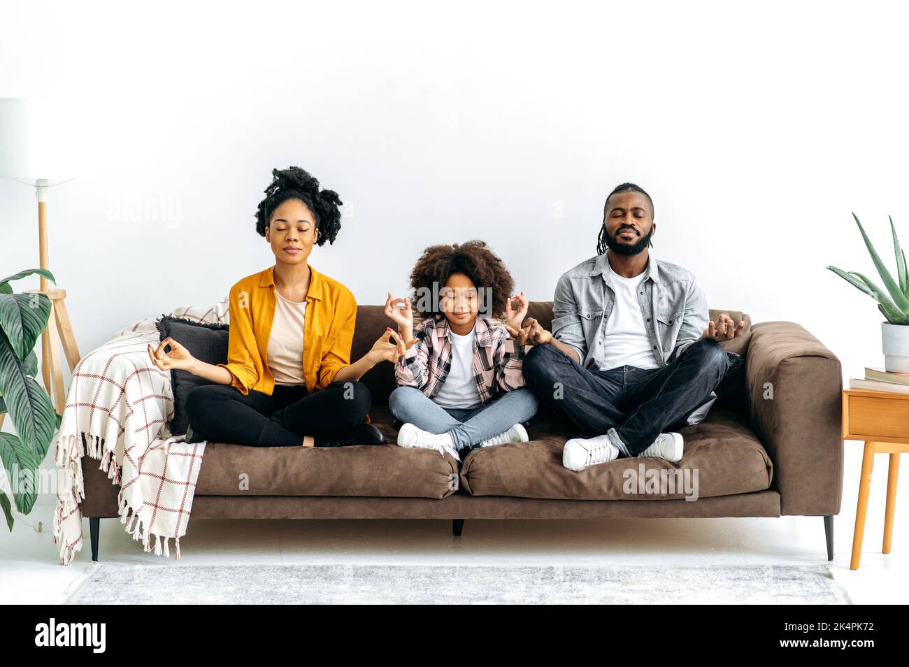 Joyeux jeune famille afro-américaine, maman et papa avec leur petite fille mignonne d'âge préscolaire, passant du temps libre à la maison ensemble, la méditation tout en étant assis sur un canapé en position lotus avec les yeux fermés, sourire Banque D'Images