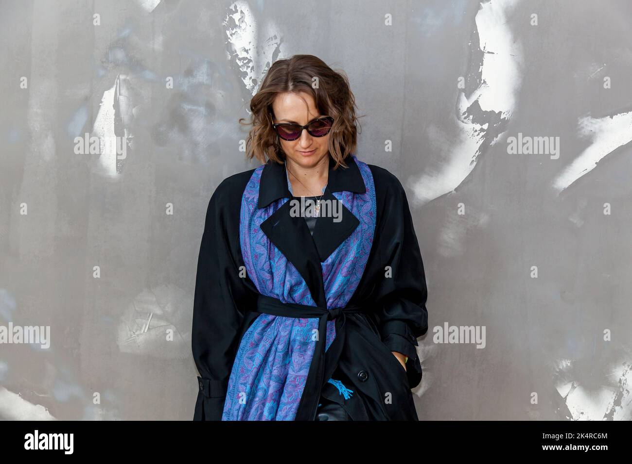 Fille tendance et élégante posant dans une robe en cuir noir, un manteau noir et des lunettes de soleil contre un mur gris Banque D'Images