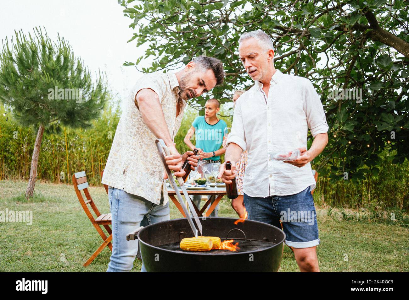 Deux hommes cuisent et toaster des bières sur un barbecue à la campagne. En arrière-plan, trois femmes amis parlent et s'amuser. Concept de style de vie. Banque D'Images