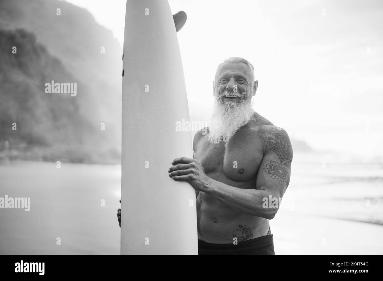 Joyeux surfeur senior tenant une planche de surf sur la plage au coucher du soleil - Focus sur le visage - montage noir et blanc. Banque D'Images