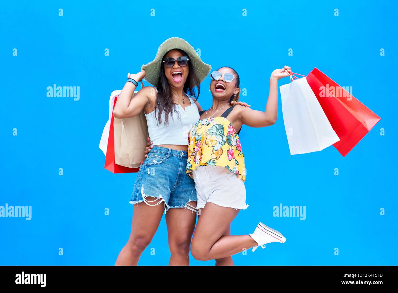 Portrait de deux jeunes femmes heureuses vêtues de vêtements d'été tenant des sacs de shopping isolés sur fond bleu, appréciant le Vendredi fou. Gaiement Banque D'Images