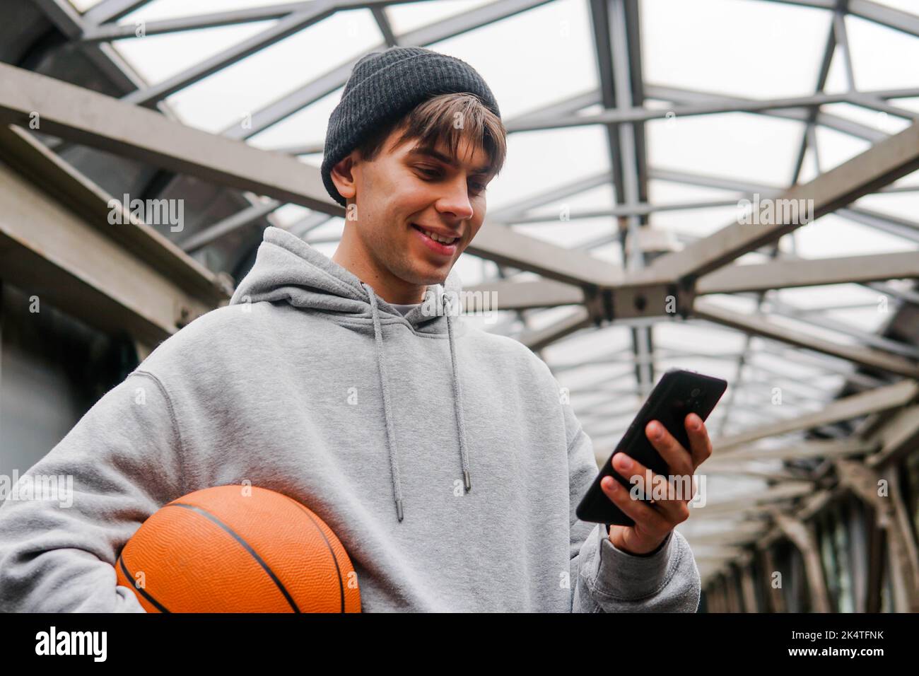 Jeune homme joueur de basket-ball avec casque tenant le ballon à l'aide d'un smartphone après l'entraînement. Fond urbain. Banque D'Images