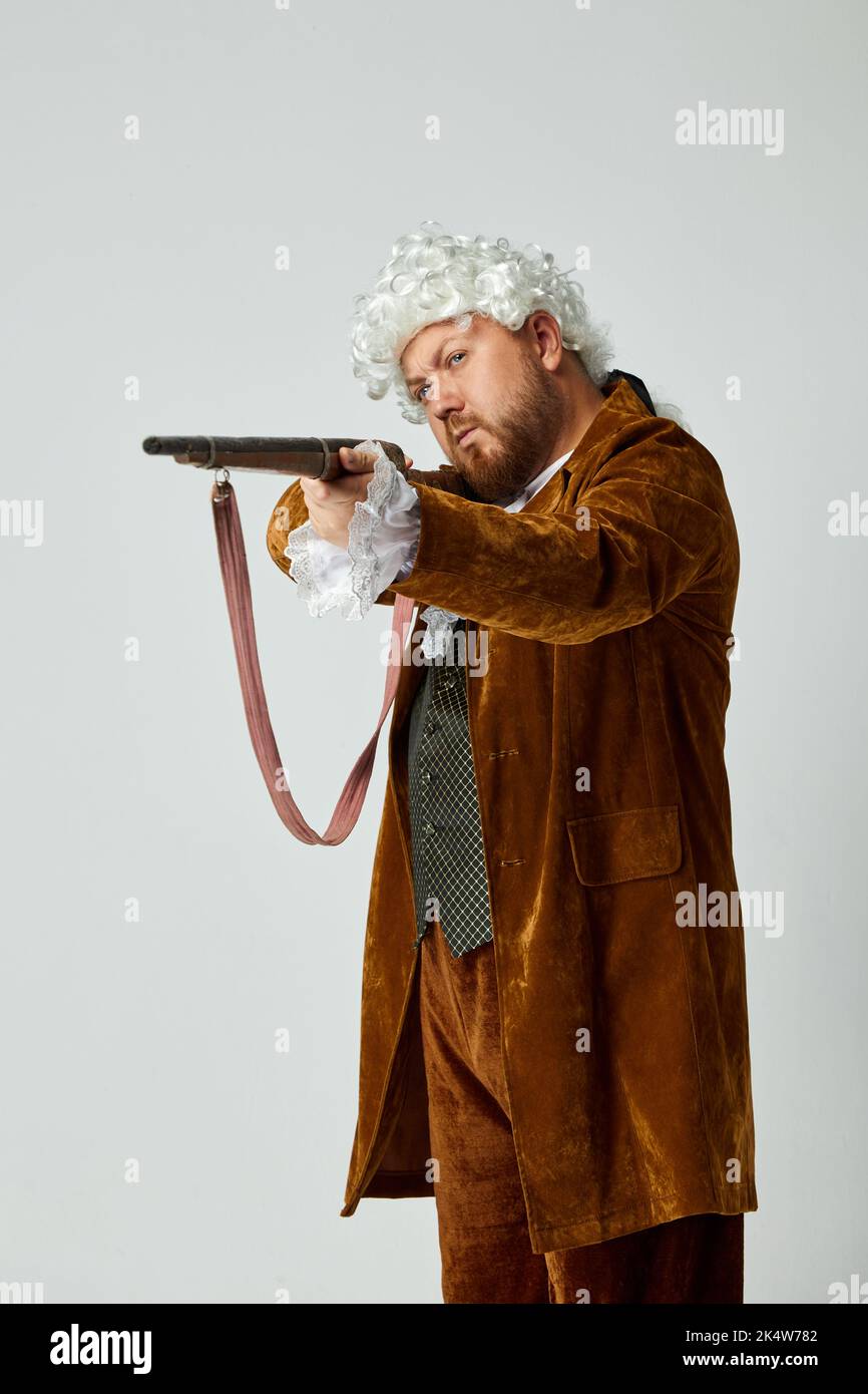 Une photo précise. Jeune homme à l'image d'une personne médiévale en costume de chasse brun vintage et perruque blanche avec ancien fusil de chasse isolé sur fond gris. Banque D'Images