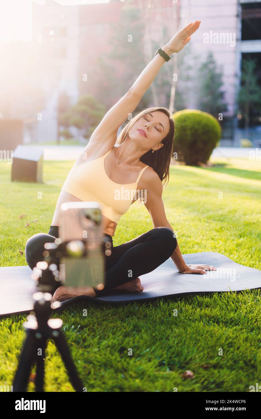 Une jeune femme blogueuse dans un sport porte en utilisant la vidéo d'enregistrement de webcam pour son blog pratiquant le yoga le matin dans le parc. Blog fitness. Sur l'herbe Banque D'Images