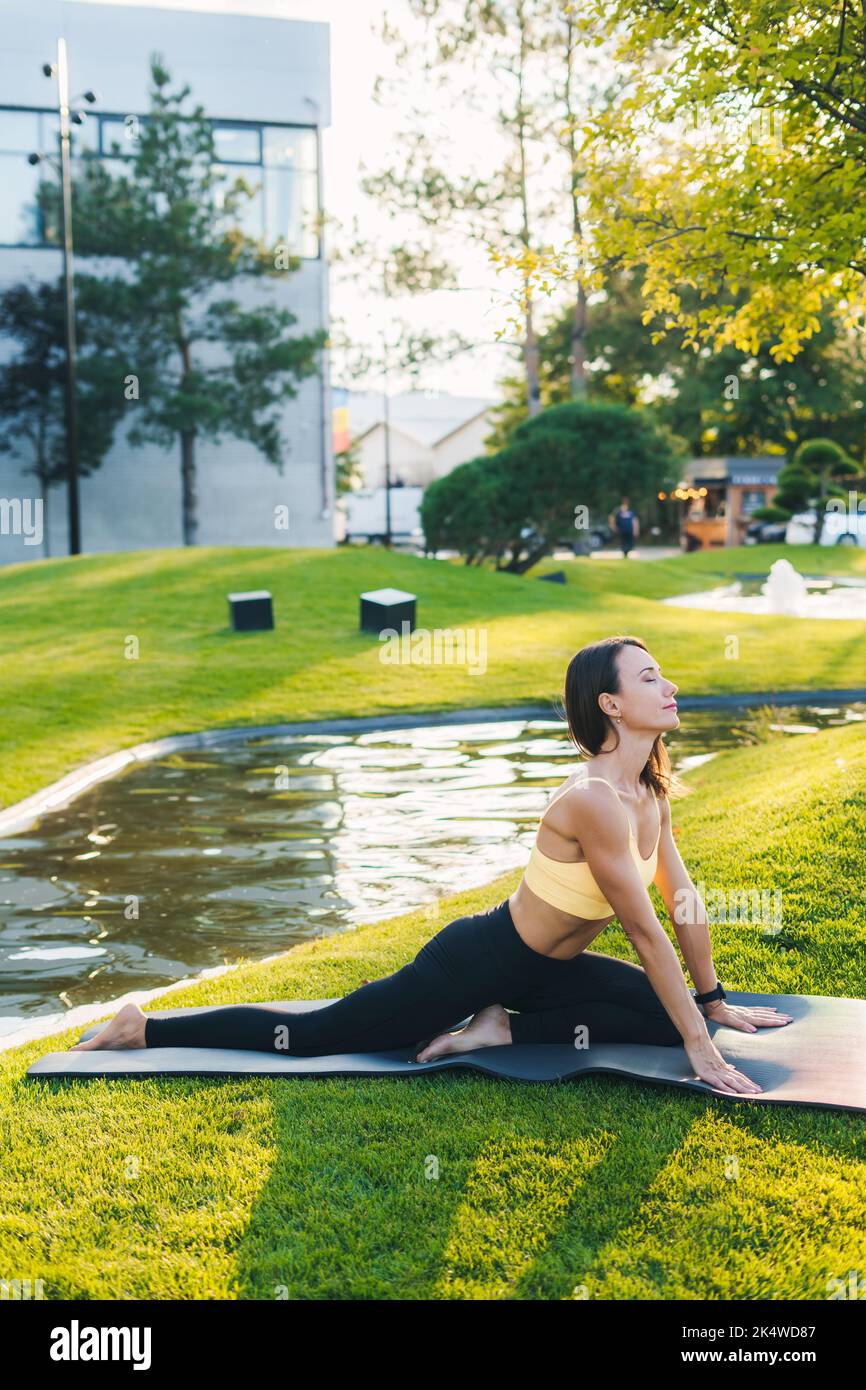 Vue latérale d'une femme athlétique assise au yoga pose sur un tapis de fitness, étirant son corps, sur un tapis dans le parc. Méditation matinale dans le parc. Plaisir Banque D'Images