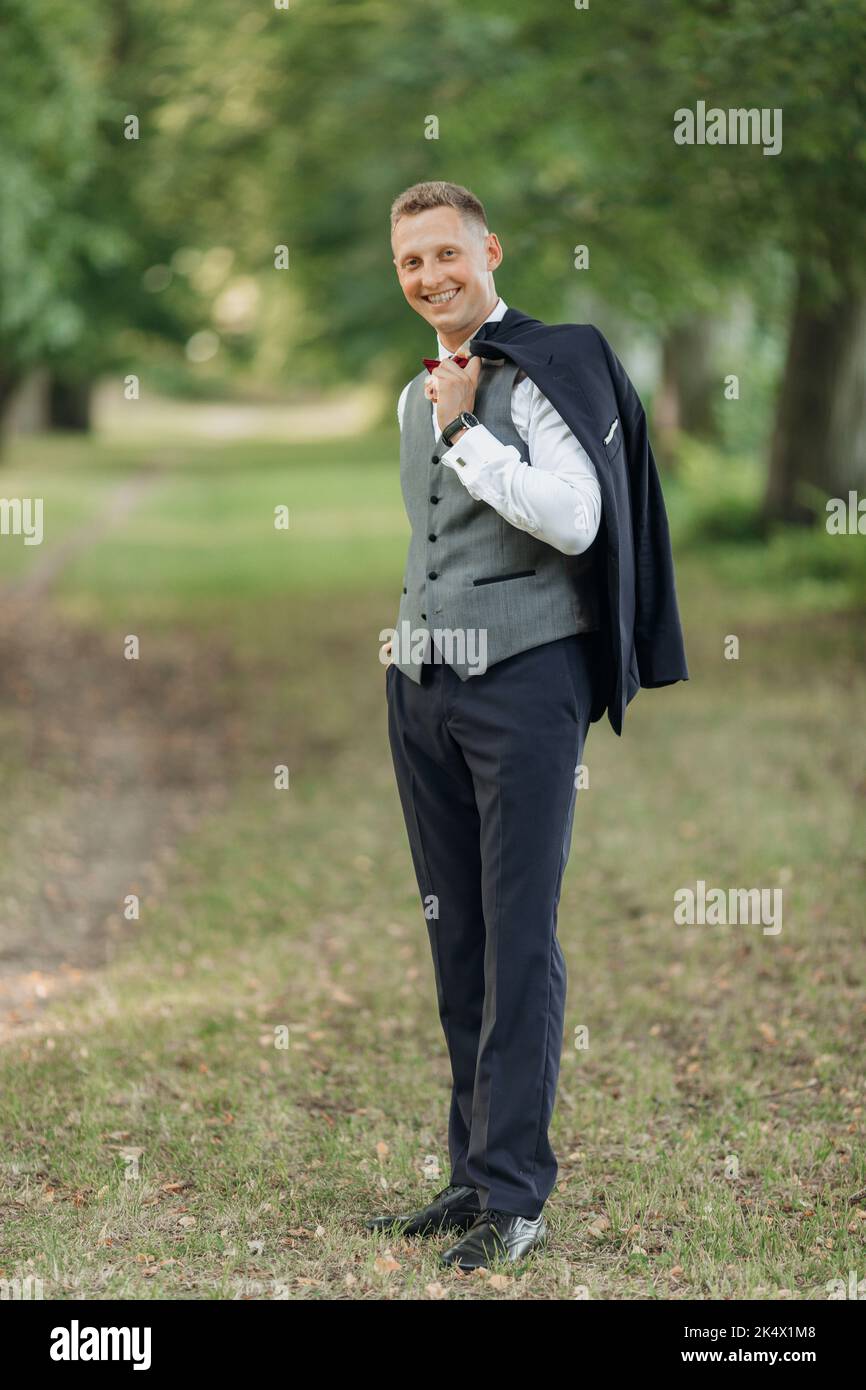 Portrait d'un jeune homme souriant marié aux cheveux courts, porter un costume bleu, un gilet gris, un noeud papillon rouge, debout au parc en été. Banque D'Images