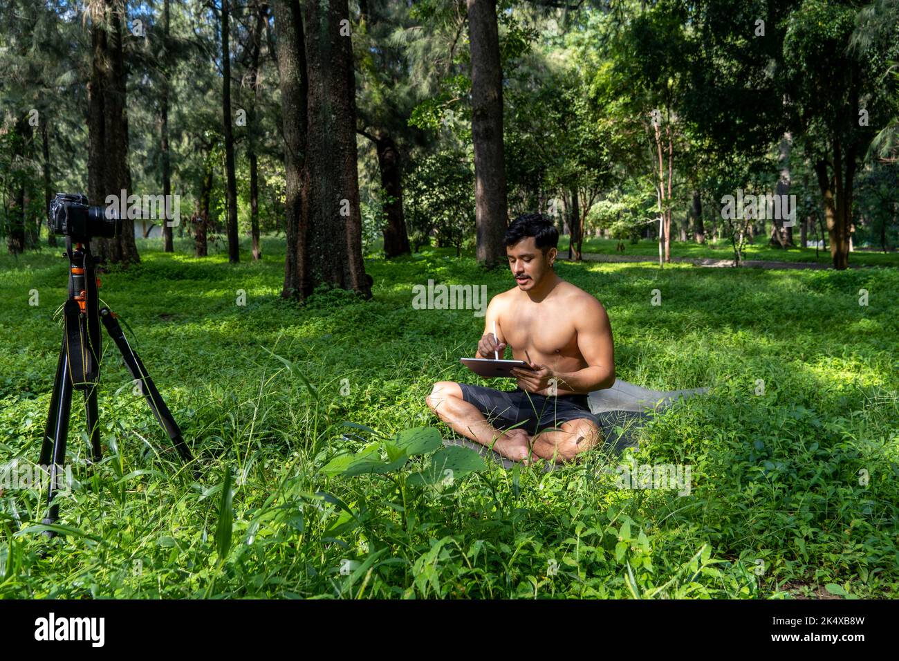 Jeune homme pratiquant le yoga asana, équilibre, méditant en se tenant sur une jambe sur tapis de sport sur l'herbe verte dans le parc. Utilisation de la tablette pour les cours en ligne. Banque D'Images
