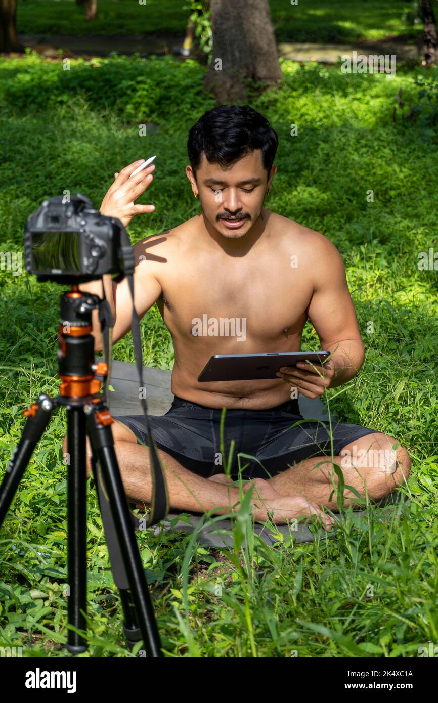 Jeune homme pratiquant le yoga asana, équilibre, méditant en se tenant sur une jambe sur tapis de sport sur l'herbe verte dans le parc. Utilisation de la tablette pour les cours en ligne. Banque D'Images