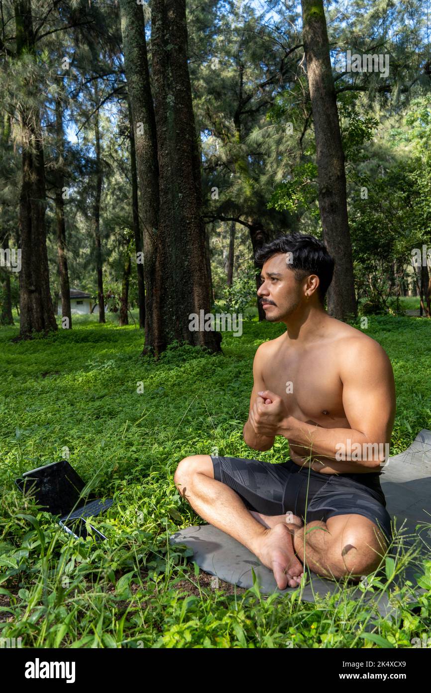 Jeune homme pratiquant le yoga asana, équilibre, méditant en se tenant sur une jambe sur tapis de sport sur l'herbe verte dans le parc. Utilisation de la tablette pour les cours en ligne. Banque D'Images