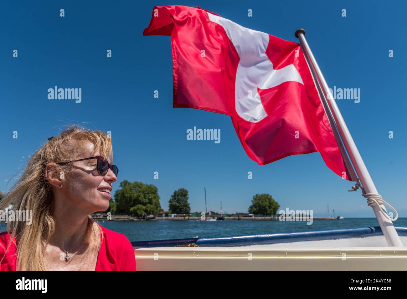 Femme blonde mature avec des lunettes de soleil souriantes et drapeau suisse flottant sur la poupe d'un bateau sur le lac Léman. Banque D'Images