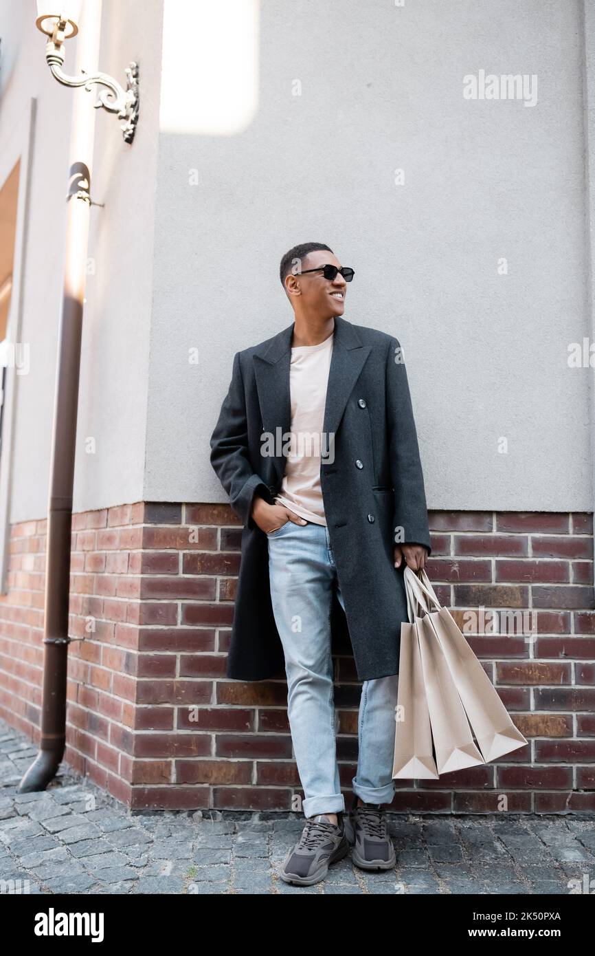 pleine longueur de l'homme afro-américain élégant en manteau et lunettes de soleil debout avec la main dans la poche des jeans et des sacs de shopping, image de stock Banque D'Images