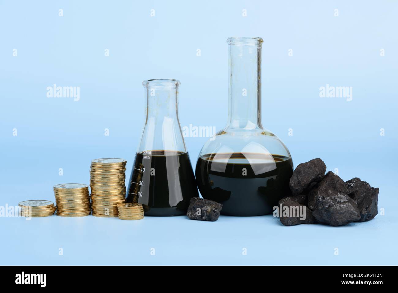 Pétrole brut dans des béchers en verre et des morceaux de charbon dur noir avec des piles de pièces sur fond bleu, concept de prix des combustibles fossiles Banque D'Images