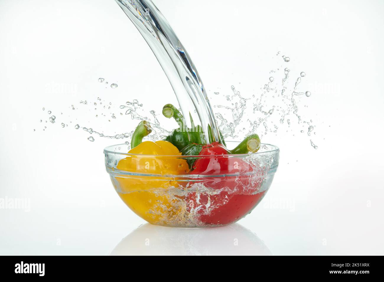 cascade qui coule sur un bol en verre avec des poivrons multicolores et des éclaboussures sur fond blanc. Banque D'Images