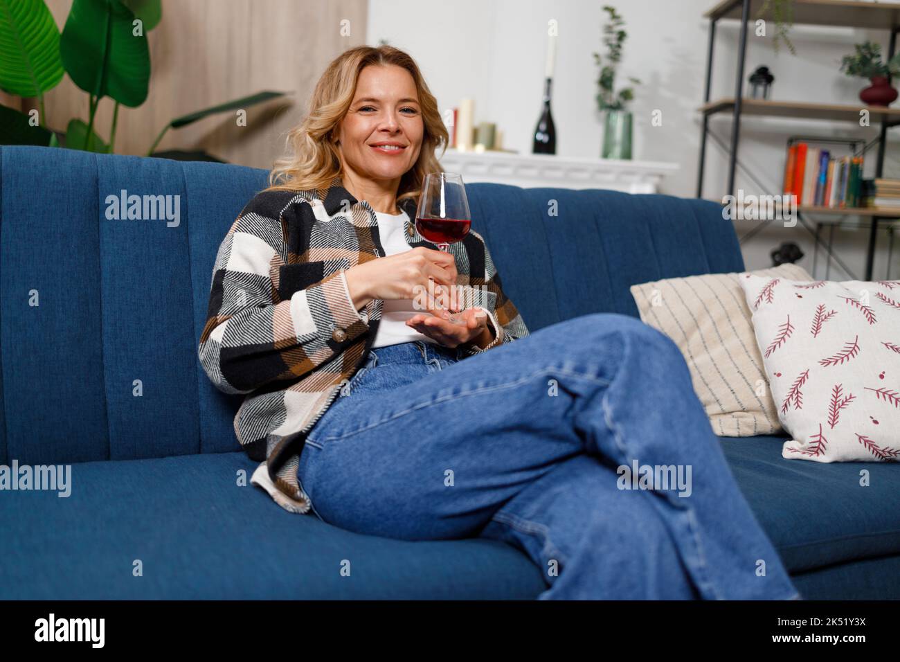 Portrait d'une jolie femme blonde d'âge moyen qui boit du vin tout en étant assise sur un canapé à la maison Banque D'Images