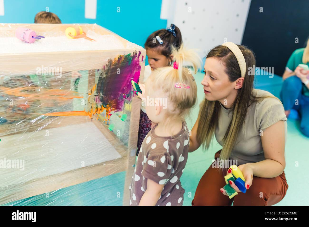 jeune et jolie enseignante aidant ses enfants dans la peinture, murs bleu de pépinière dans le fond. Photo de haute qualité Banque D'Images