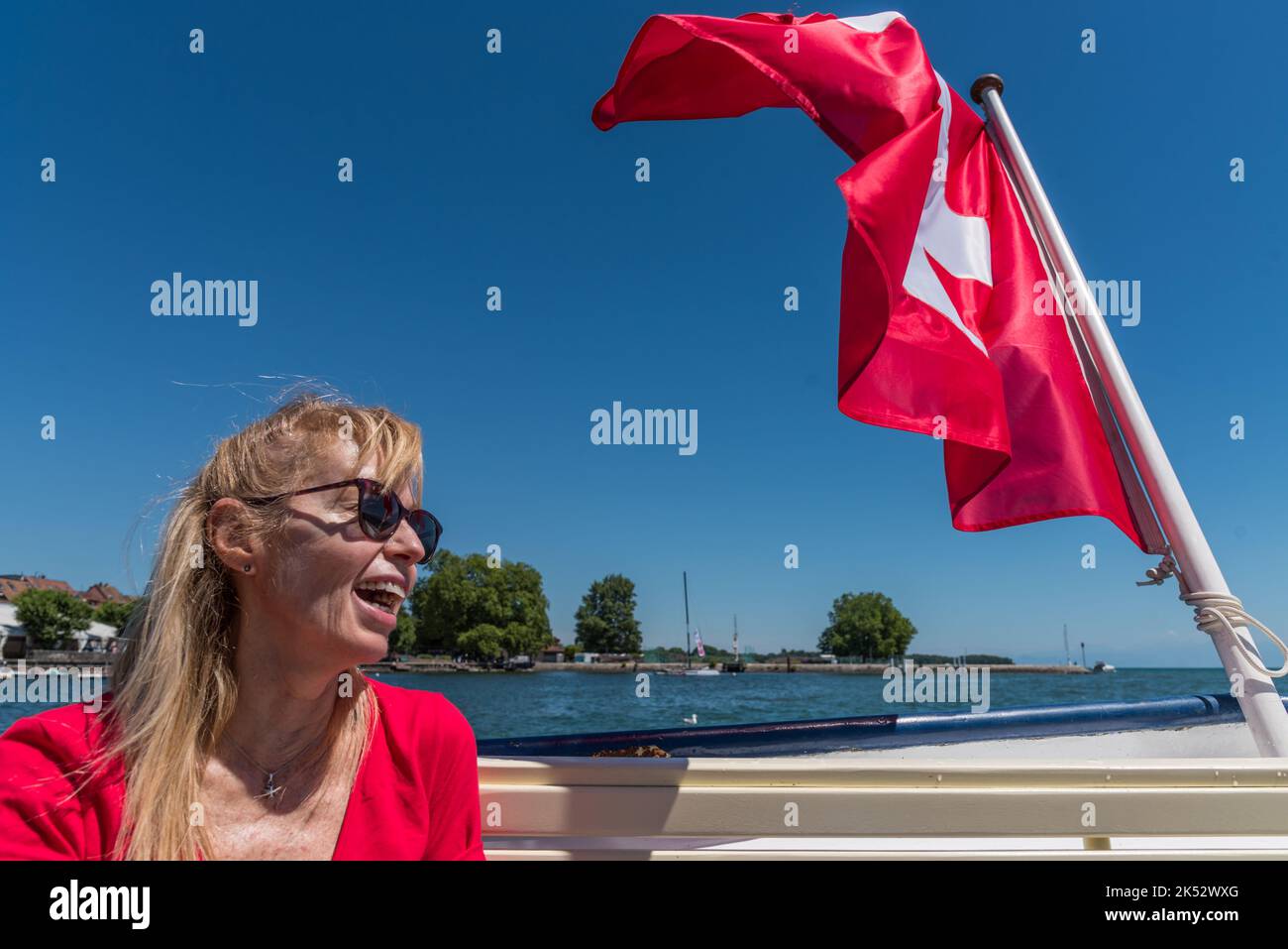 Femme blonde mûre avec des lunettes riant et drapeau suisse flottant à l'arrière d'un bateau sur le lac Léman. Banque D'Images