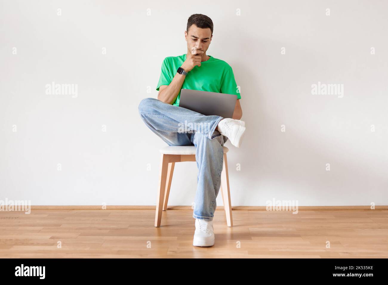 Homme arabe pensif utilisant un ordinateur portable et pensant, naviguant sur Internet tout en étant assis sur une chaise contre le mur blanc à la maison Banque D'Images
