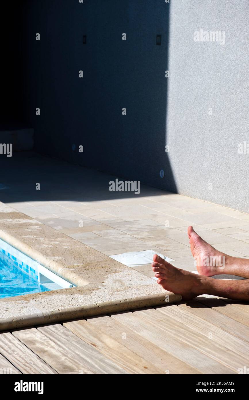 Les pieds mâles sur le bord d'une piscine avec l'homme anonyme se détendant au soleil. Banque D'Images
