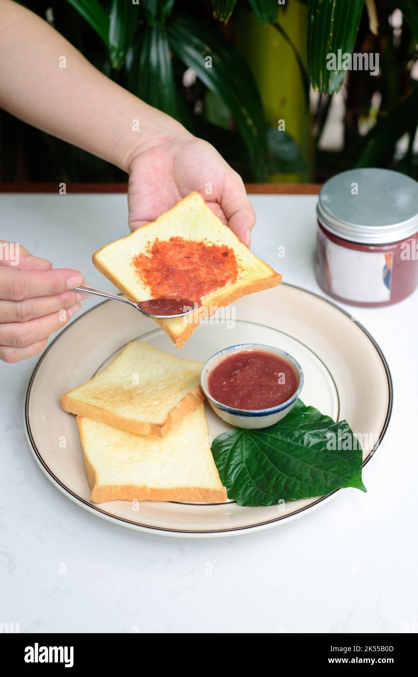 Les mains répandant la confiture de prune sur un toast contre le pot avec la confiture et les fleurs d'hibiscus Banque D'Images