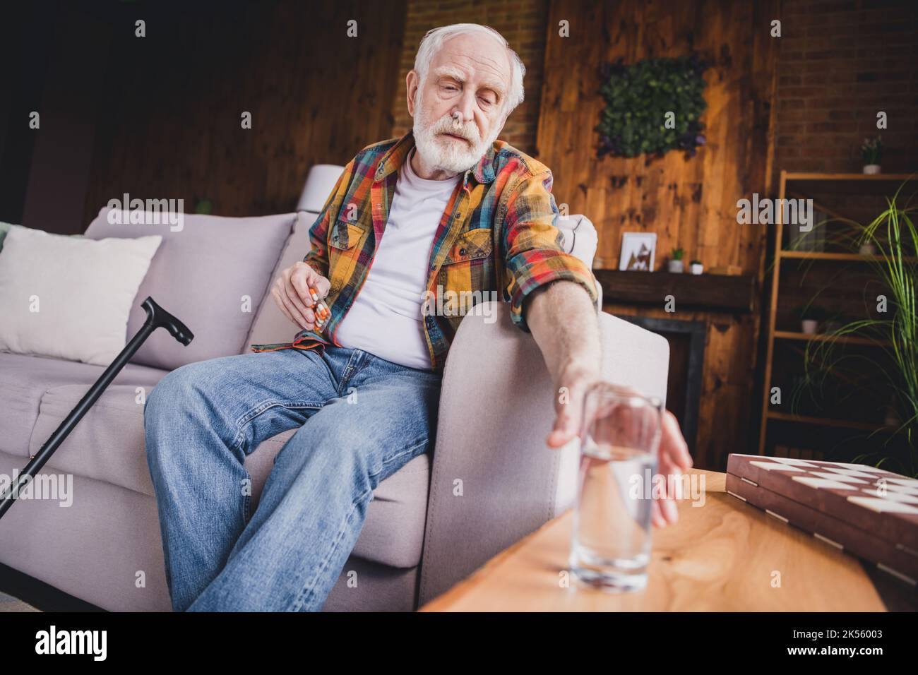 Photo de solitaire bouleversé retraité homme habillé chemise de plaid assis canapé buvant des pilules d'eau à l'intérieur de l'appartement Banque D'Images