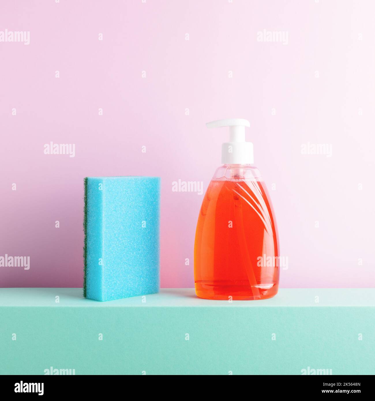 Composition esthétique et colorée de vaisselle carrée avec éponge de nettoyage et savon dans une bouteille debout sur un podium sur fond rose. Banque D'Images