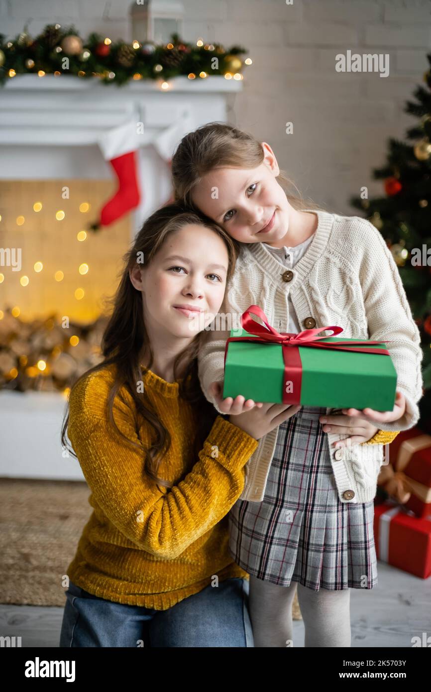 sœurs souriantes tenant une boîte cadeau et regardant l'appareil photo dans le salon avec décoration de noël, image de stock Banque D'Images