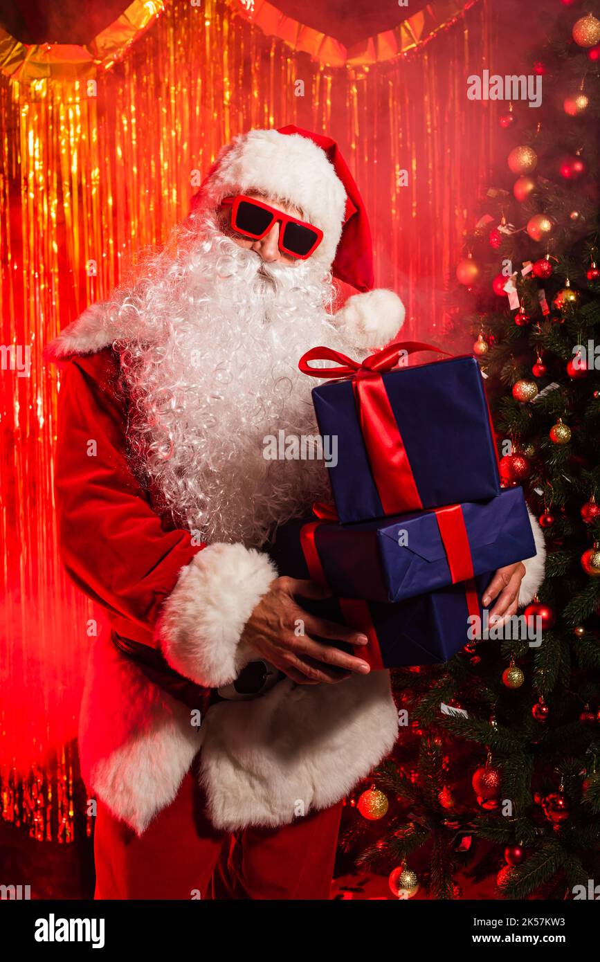 Le Père noël en lunettes de soleil tenant des cadeaux près de l'arbre de noël et scintillant, image de stock Banque D'Images