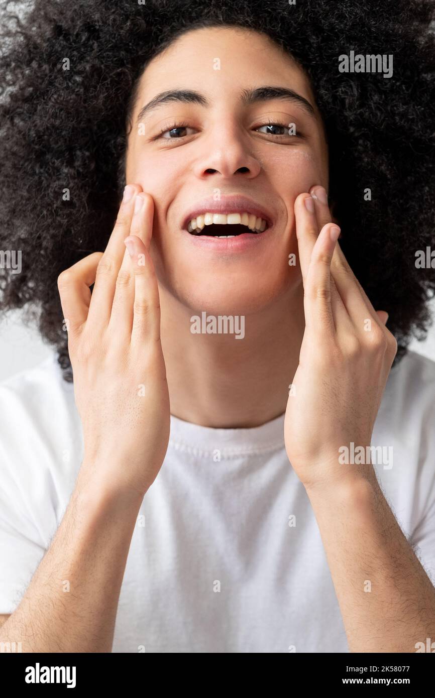 Soin du visage pour homme. Traitement de la peau. Traitement de l'acné. Joyeux satisfait gars beau avec les cheveux longs bruns bouclés dans le t-shirt blanc appliquant la crème appréciant touc Banque D'Images Soin du visage pour homme. Traitement de la peau. Traitement de l'acné. Joyeux satisfait gars beau avec les cheveux longs bruns bouclés dans le t-shirt blanc appliquant la crème appréciant touc Banque D'Images